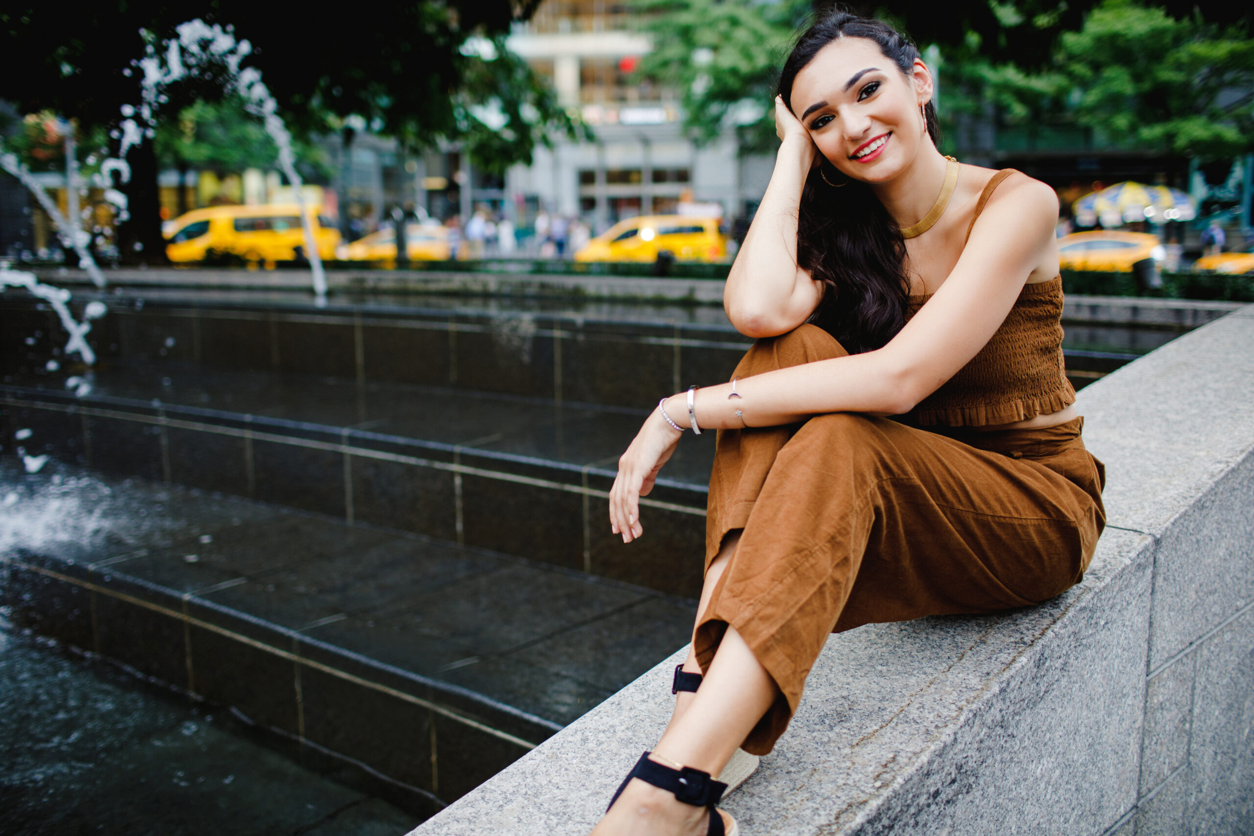 Brunetter Senior Model poses in a trendy brown two-piece set and black strappy heels with one hand in her hair sitting on a concrete wall in New York City with Yellow Taxis in the background 
