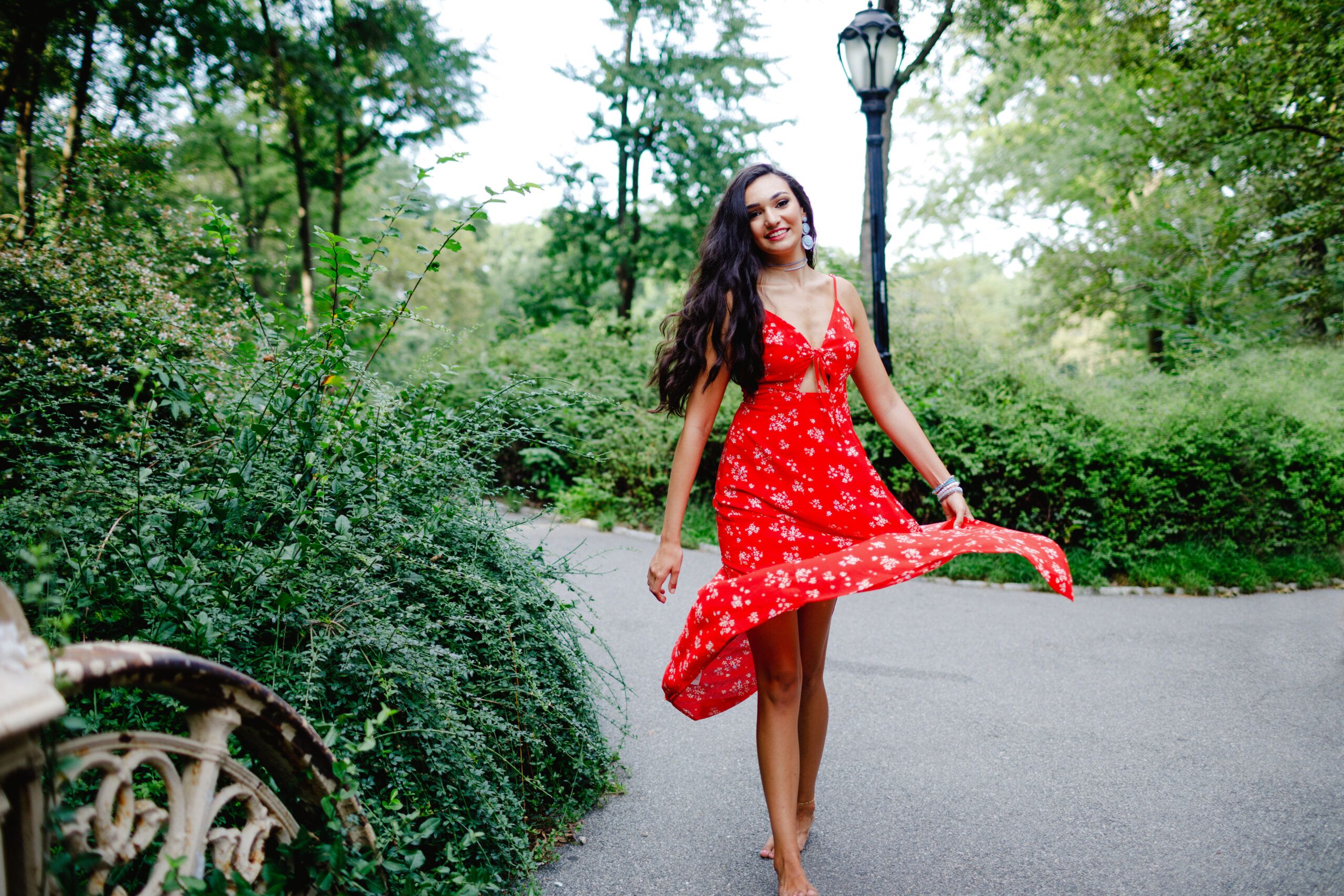 Senior Photo of a Brunette Model with long hair and red lipstick posing in Central Park in a red floral dress walking