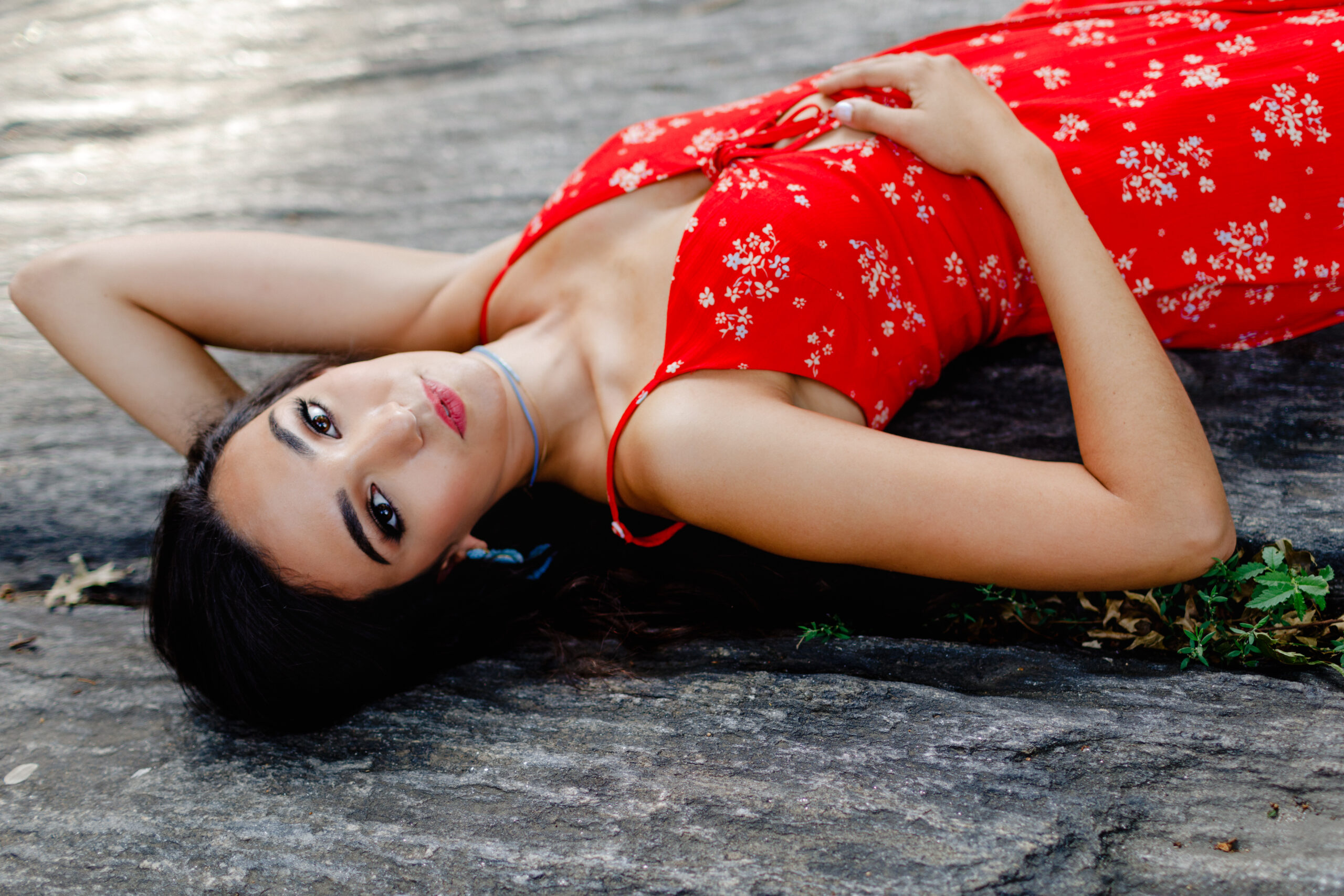Senior Photo of a Brunette Model with long hair and red lipstick posing in Central Park in a red floral dress laying on a rock