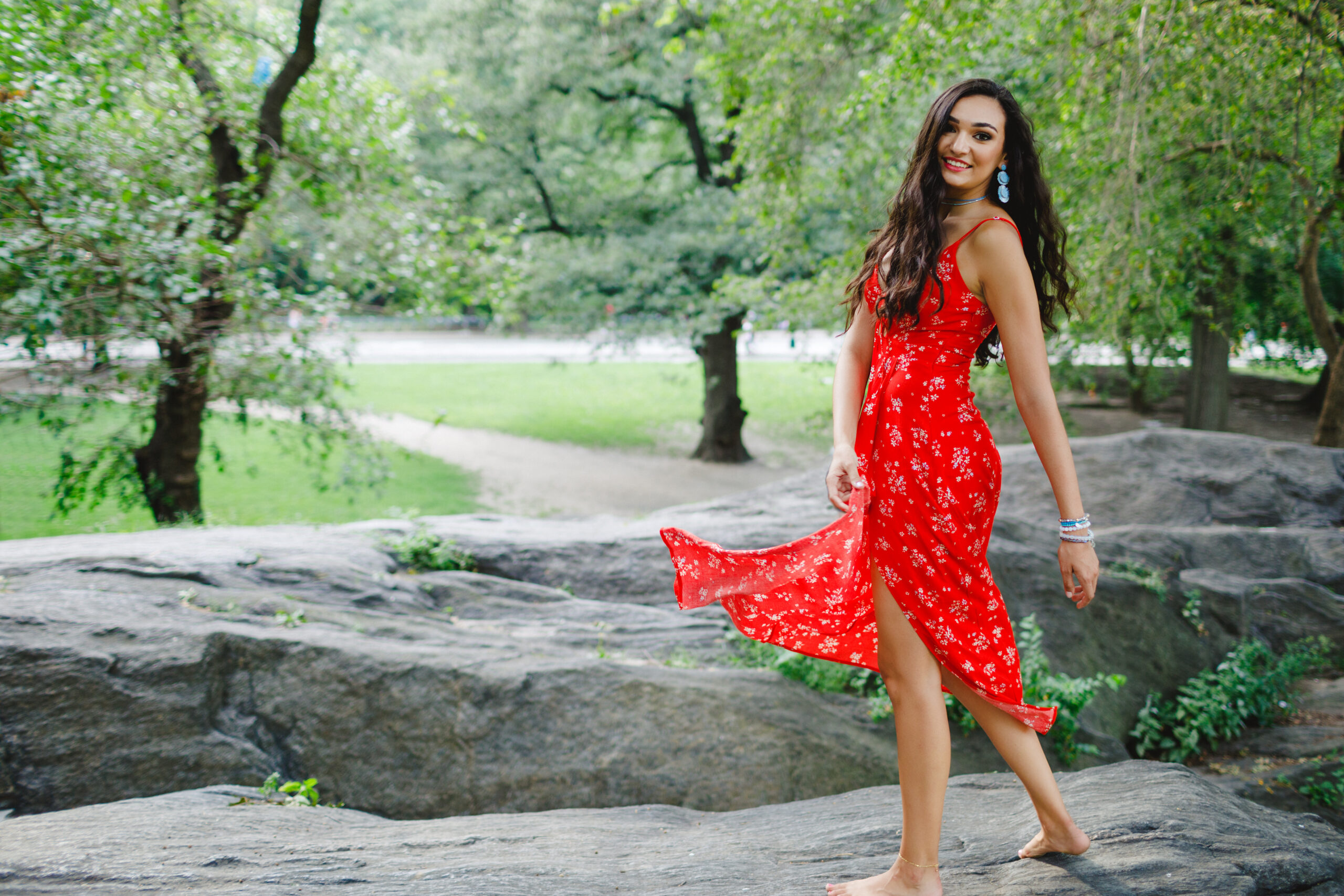 Senior Photo of a Brunette Model with long hair and red lipstick posing in Central Park in a red floral dress twirling her dress standing on a large rock