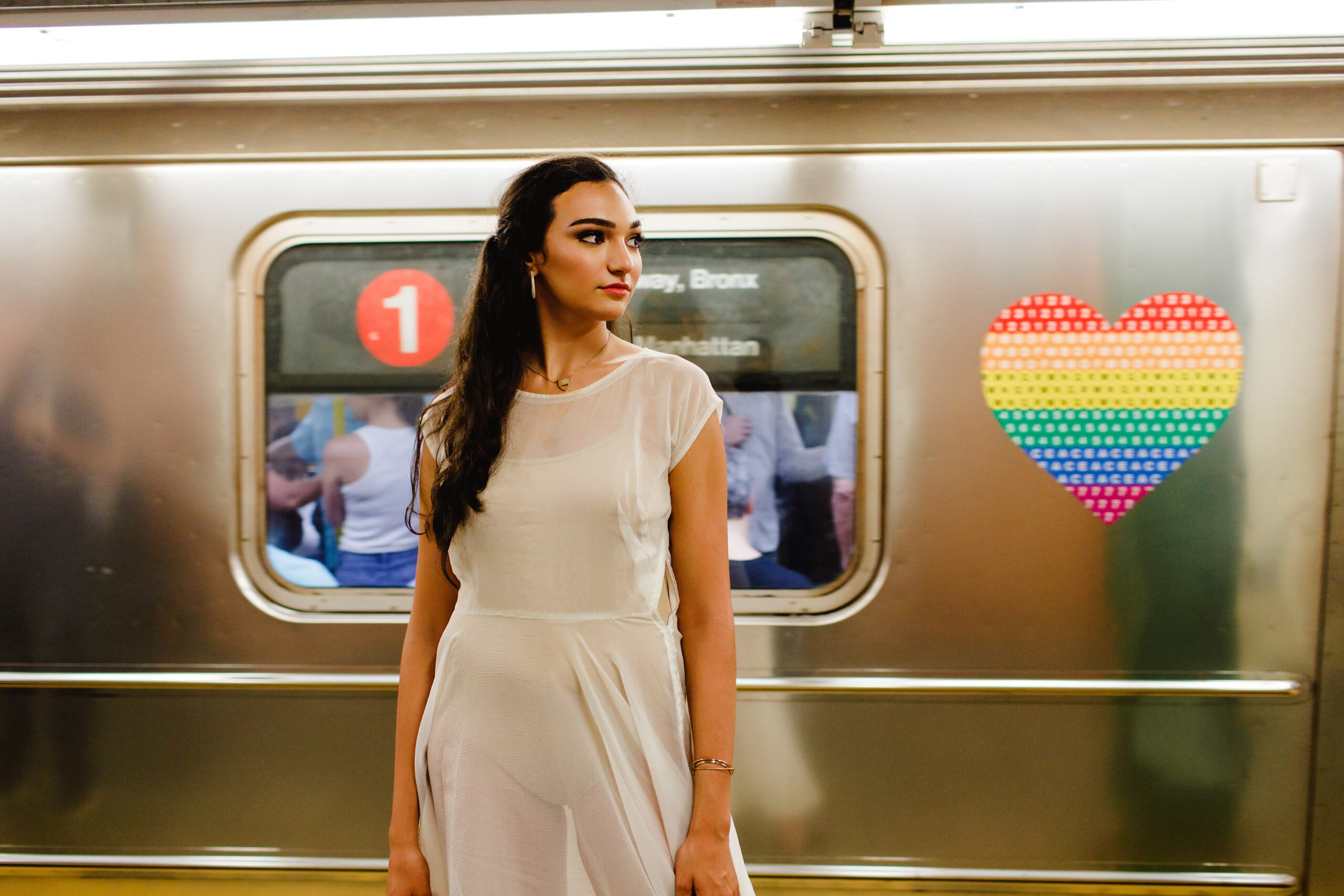 Girl with long dark hair and red lips in a white dress staring off into the distance while standing beside a moving subway car with a rainbow heart on the outside of it. 