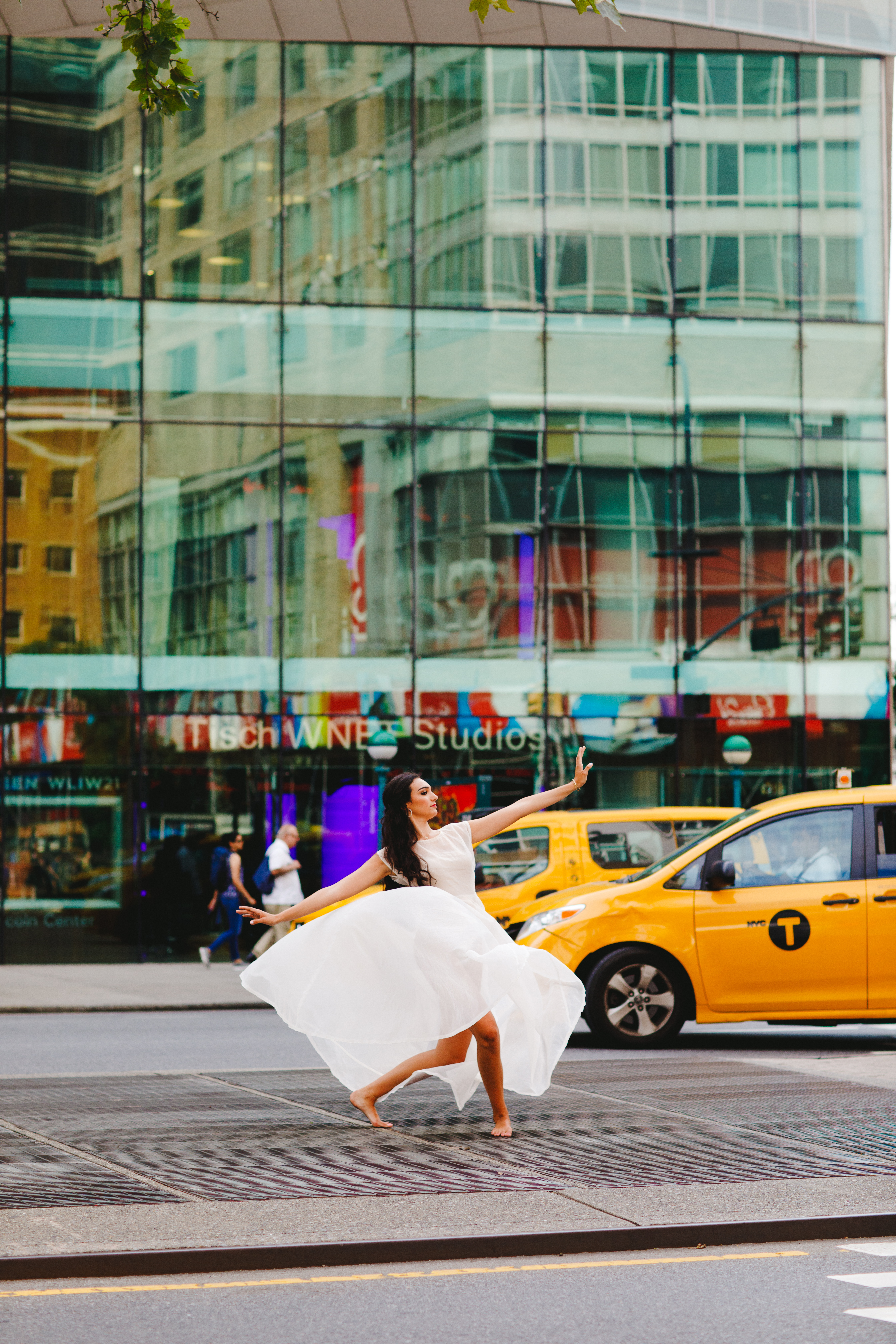 Brunette girl with long hair and red lipstick wearing a white dress and pink satin pointe shoes in a lunge on the streets of NYC in front of yellow taxis.