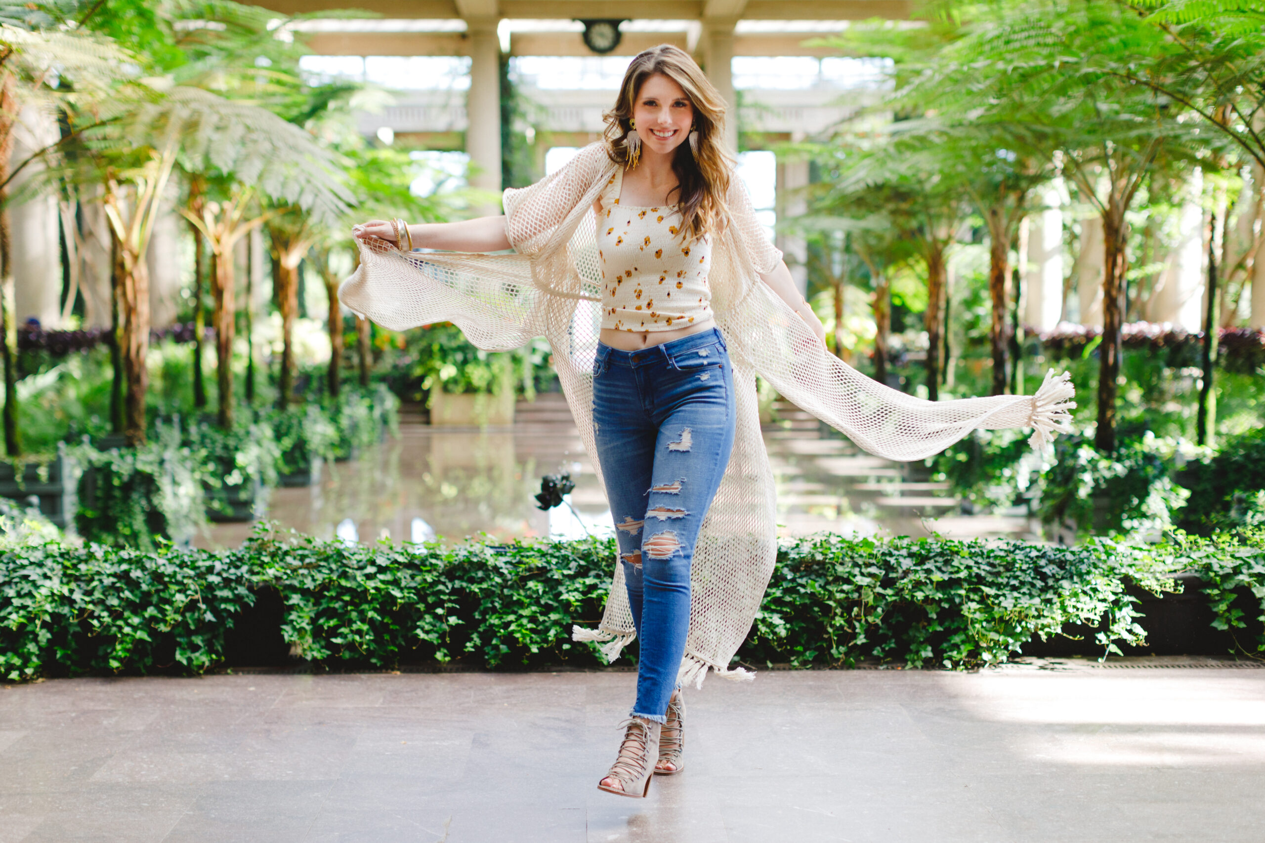 Clare throwing a white netting kimono behind her in jeans and a white floral tank top in the greenhouse at Longwood gardens.