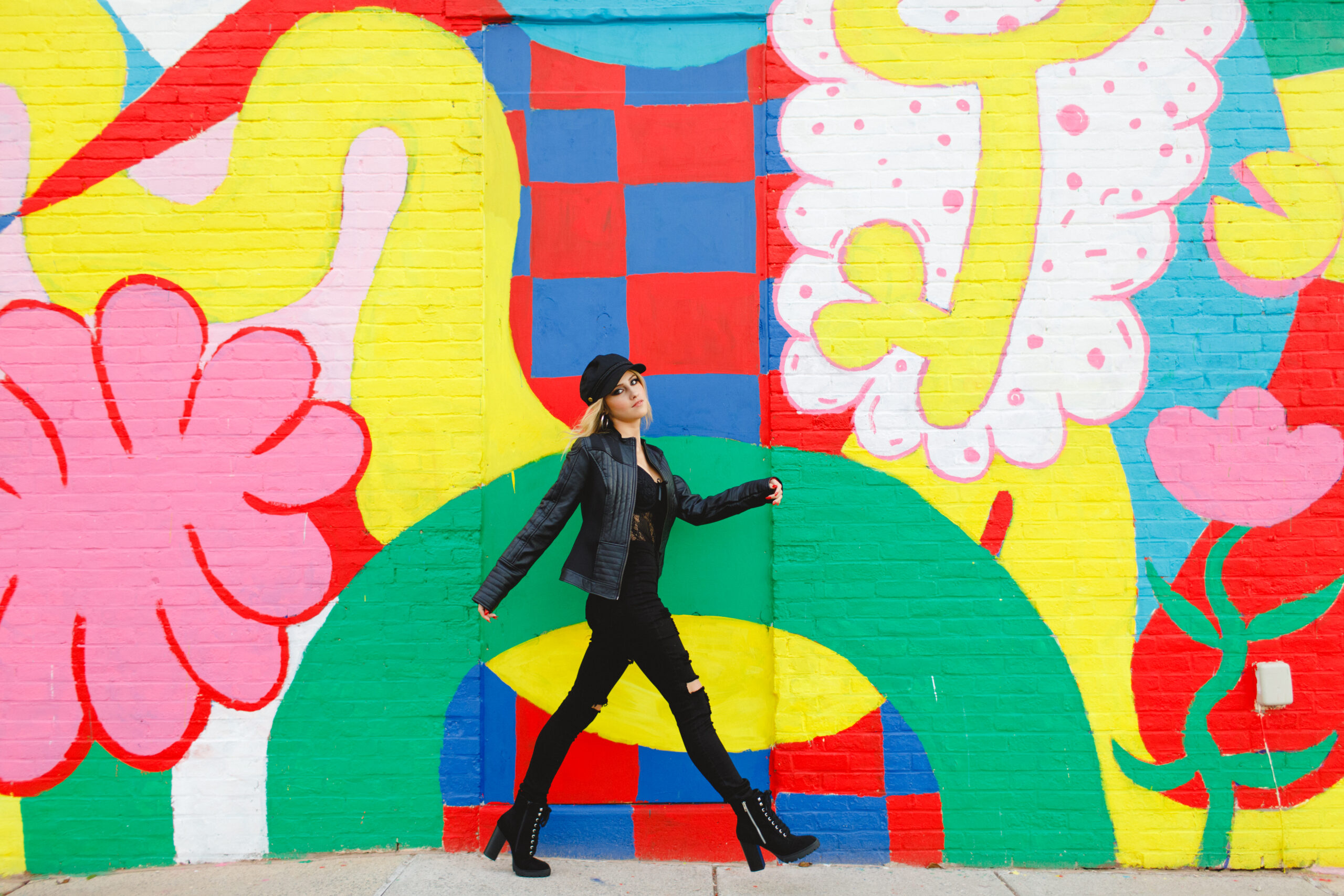 Andrina walking down the sidewalk in an all black outfit in front of a bright colored trendy mural
