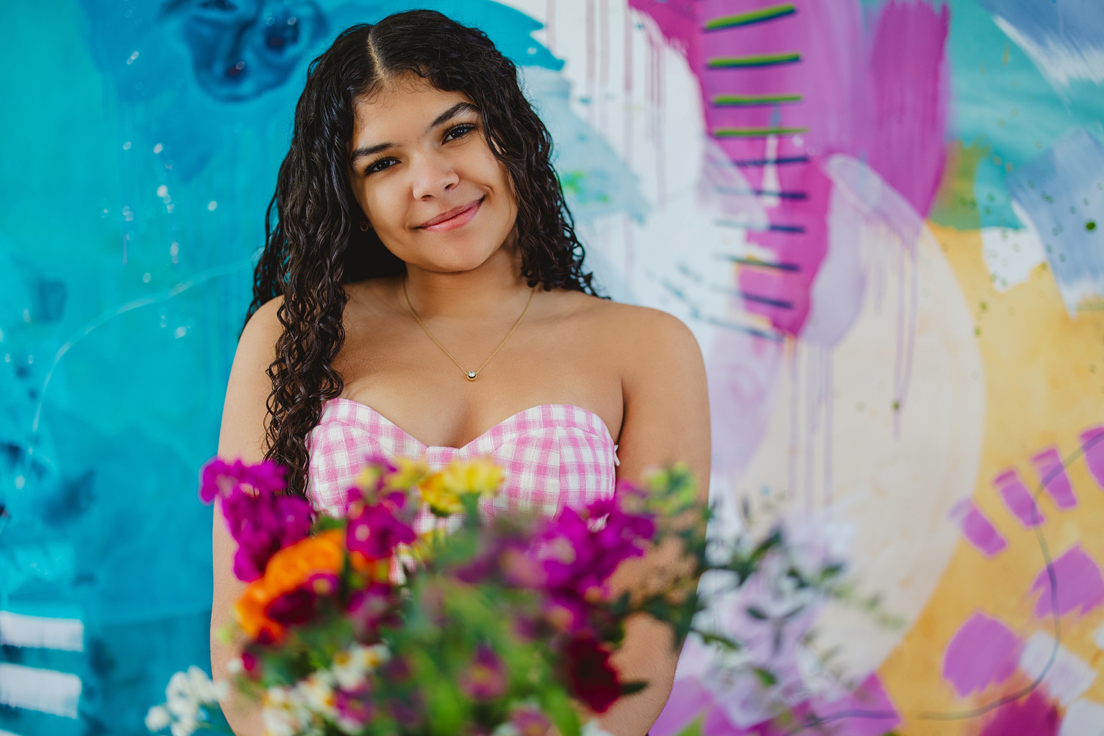 Biracial girl with long curly hair wearing a gingham tube top holding a bouquet of flowers in front of vibrant mural 